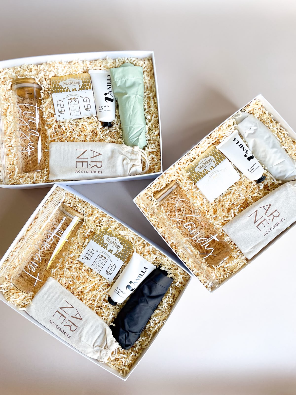 Three white open gift boxes with cream crinkle paper each containing an engraved glass tumbler, MaMere nougat, M'Beauty Vanilla hand cream, a ZARNÉ accessories pouch and a folded umbrella