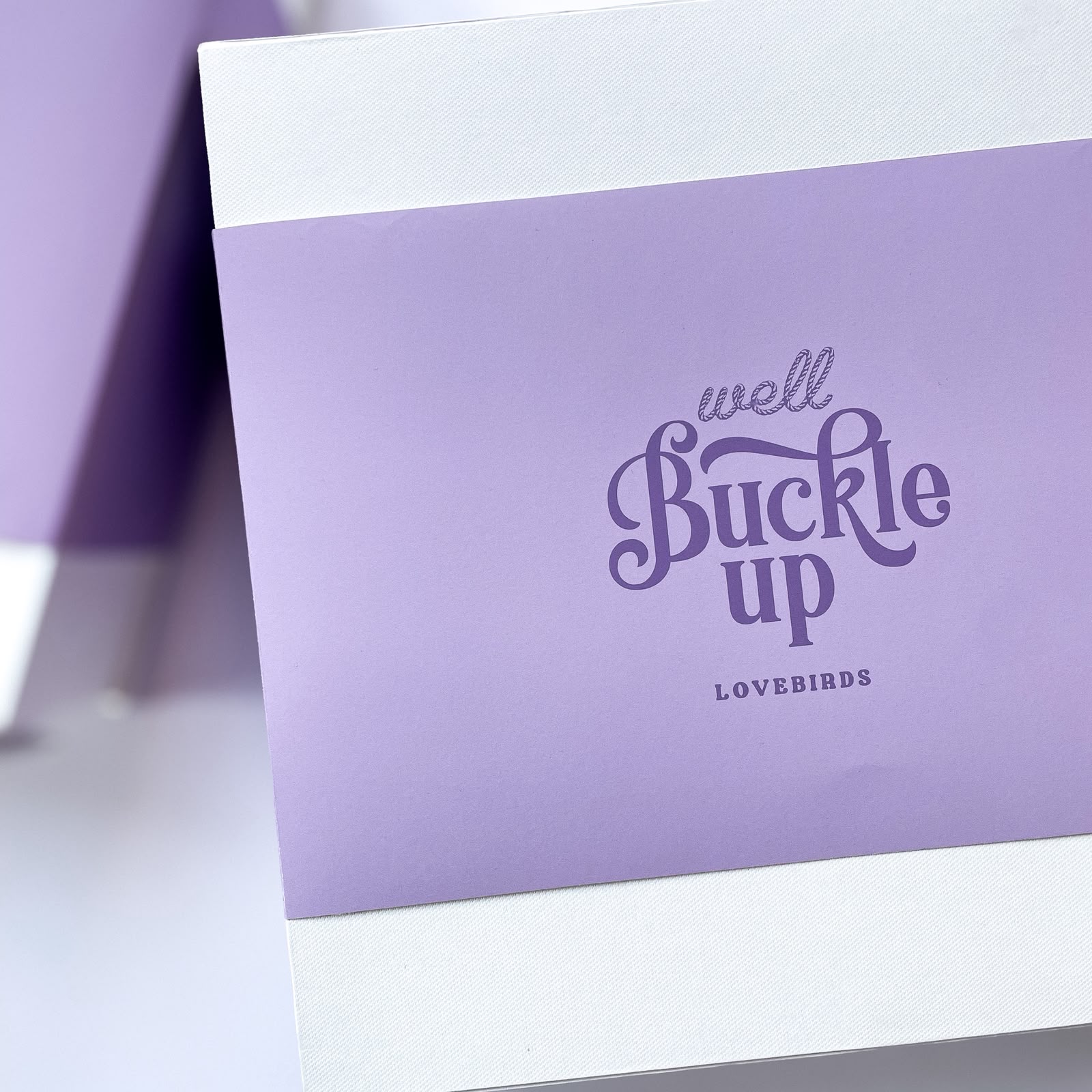 Close-up of a lavender gift box with a purple wrap-around band reading 'Well Buckle Up Lovebirds' in elegant script
