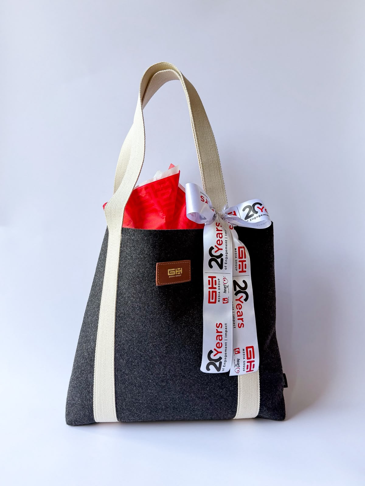 Dark grey GH Media canvas tote bag with cream handles tied with a branded '20 Years of Engagement' ribbon and filled with red tissue paper