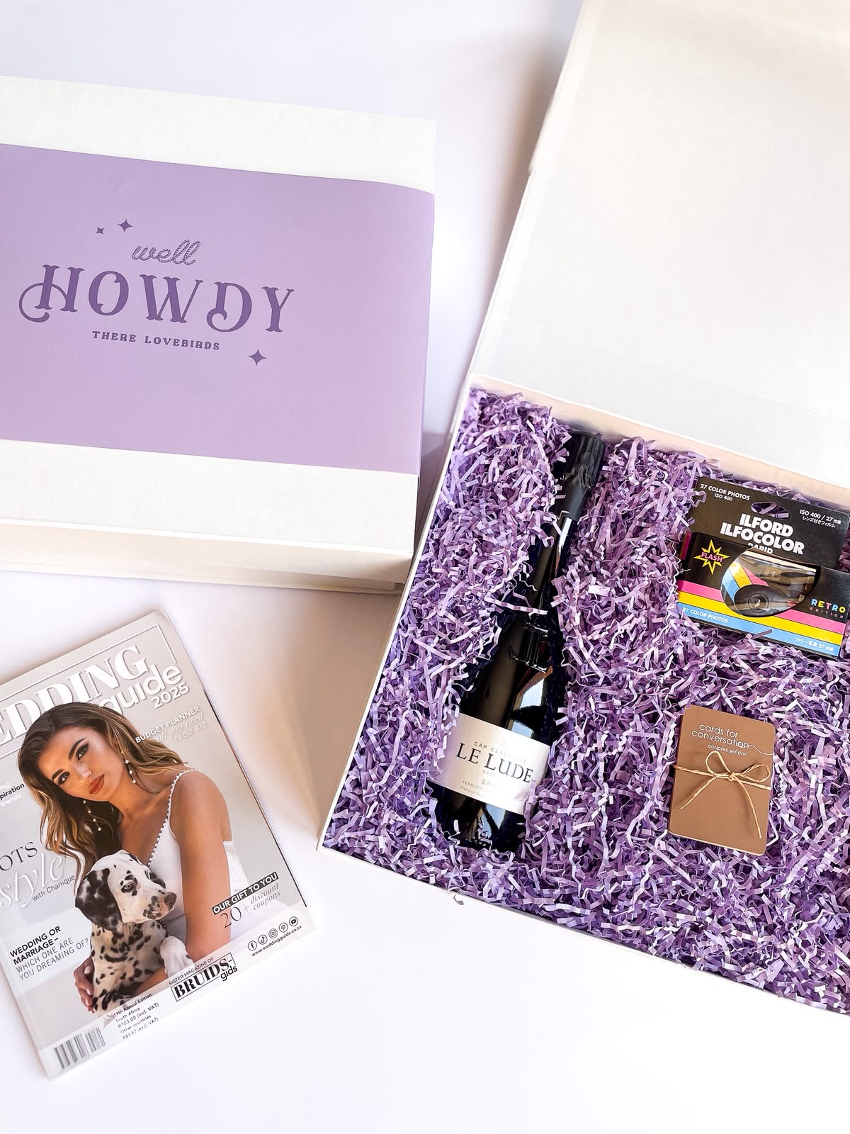 White gift box with purple lid reading 'Well Howdy There Lovebirds' open to reveal purple crinkle paper with a Le Lude sparkling wine, Ilford film roll and Cards for Conversation booklet