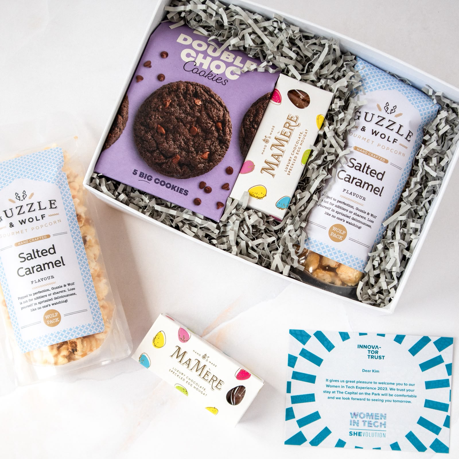 Open white gift box with grey crinkle paper containing Double Choc Cookies, MaMere nougat and Guzzle & Wolf Salted Caramel popcorn beside an Innovator Trust Women in Tech SHEvolution branded card