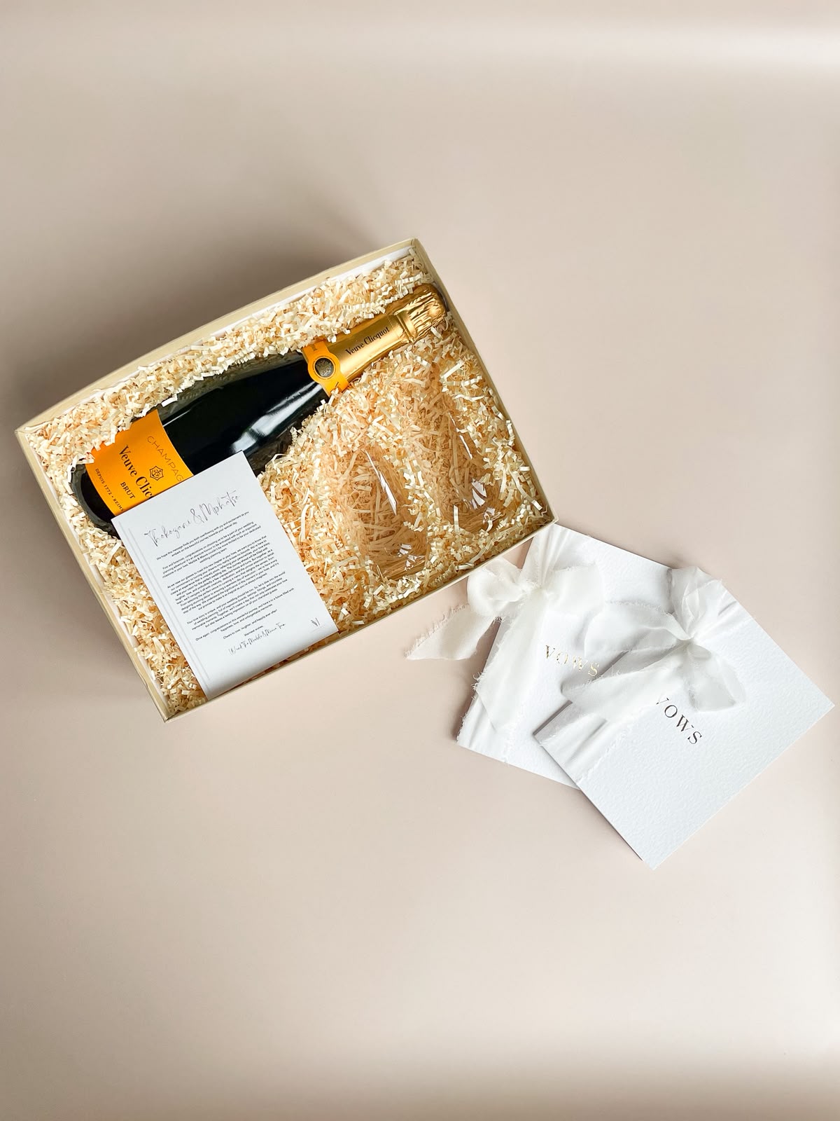 Open cream gift box with wood-shred crinkle paper containing a Veuve Clicquot champagne bottle, a personalised welcome letter and an embossed white vows booklet with satin bow
