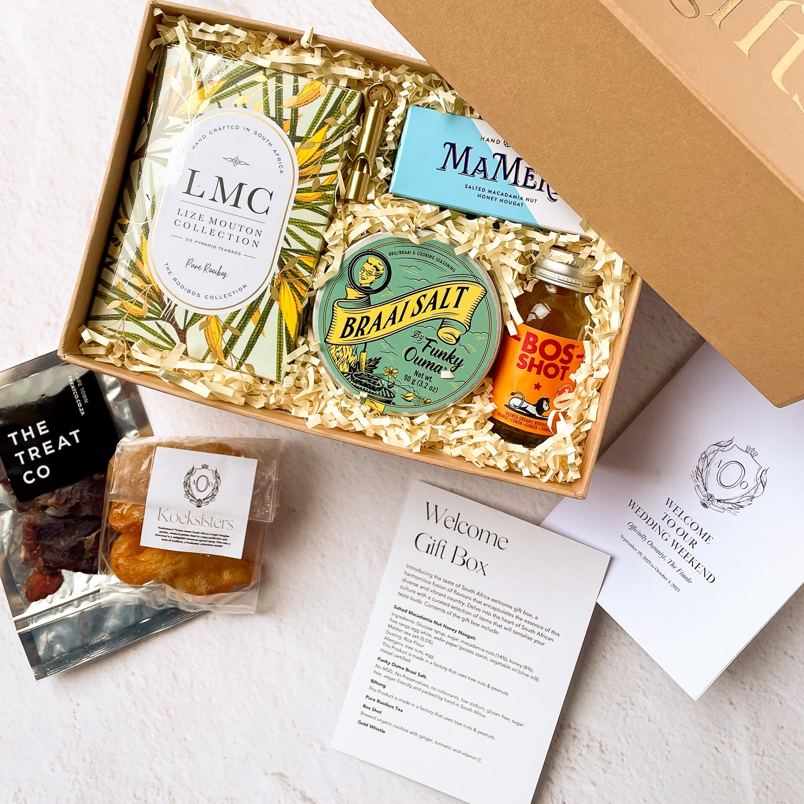 Open kraft gift box with cream crinkle paper containing LMC Rooibos tea, Braai Salt by Funky Ouma, BosShot, MaMere nougat and The Treat Co koeksisters beside a Welcome to Our Wedding Weekend card